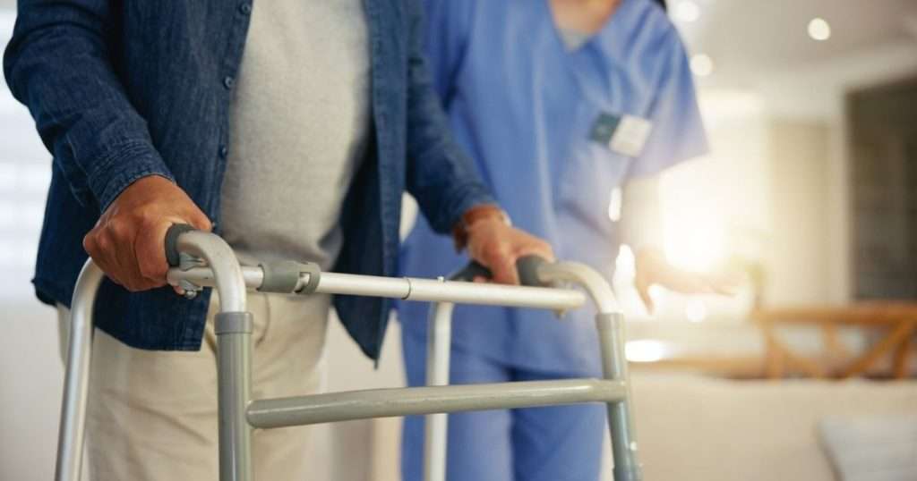 An older man using a walker while assisted by a healthcare professional during physical therapy or rehabilitation is at an increased risk of fall-related injuries and bedsores due to reduced mobility
