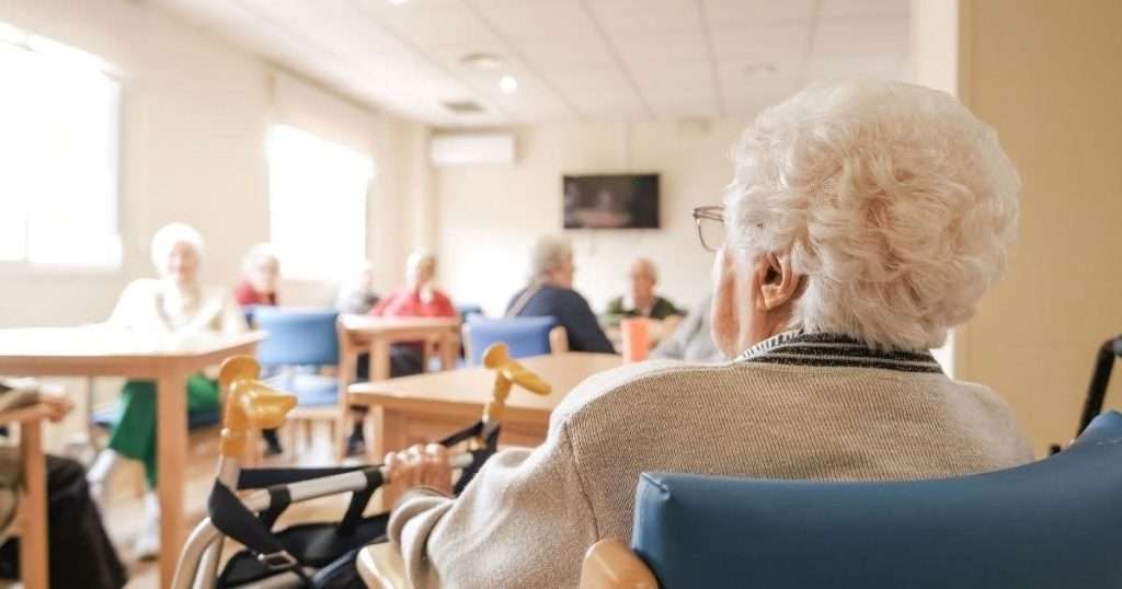 Elderly woman with a foldable walker sitting alone in an activities room in a skilled nursing facility or assisted living facility with other older adults in the background